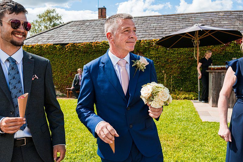 A man in a blue suit holds a bouquet of white flowers while standing outdoors on a sunny day, accompanied by others in formal attire. Image by Documentary Wedding Photographer Lewis Bishop Photography.
