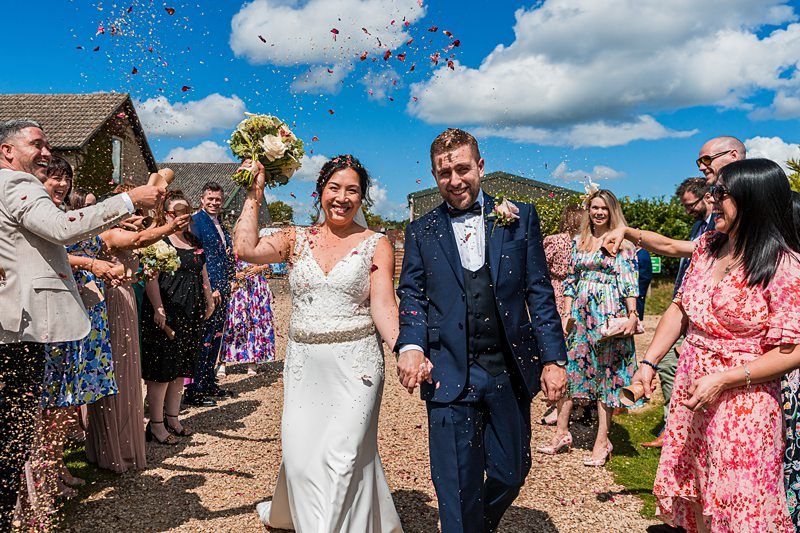 A bride and groom walk hand-in-hand outdoors, smiling as wedding guests throw confetti. The sky is blue with scattered clouds, and guests are dressed in colorful attire. Image by Documentary Wedding Photographer Lewis Bishop Photography.