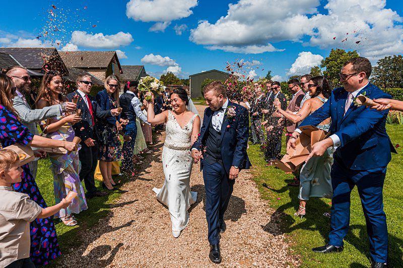 A bride and groom walk hand in hand surrounded by guests throwing confetti, on a sunny day outside. Image by Documentary Wedding Photographer Lewis Bishop Photography.