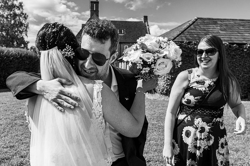 A man in sunglasses hugs a bride holding a bouquet, while a smiling woman in sunglasses stands nearby outdoors on a sunny day. Image by Documentary Wedding Photographer Lewis Bishop Photography.