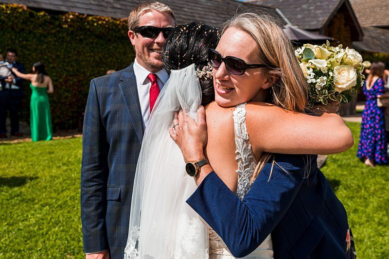 A bride in a white dress and veil is hugging a woman holding a bouquet while a man in a suit and sunglasses stands nearby on a grassy area with other people in the background. Image by Documentary Wedding Photographer Lewis Bishop Photography.