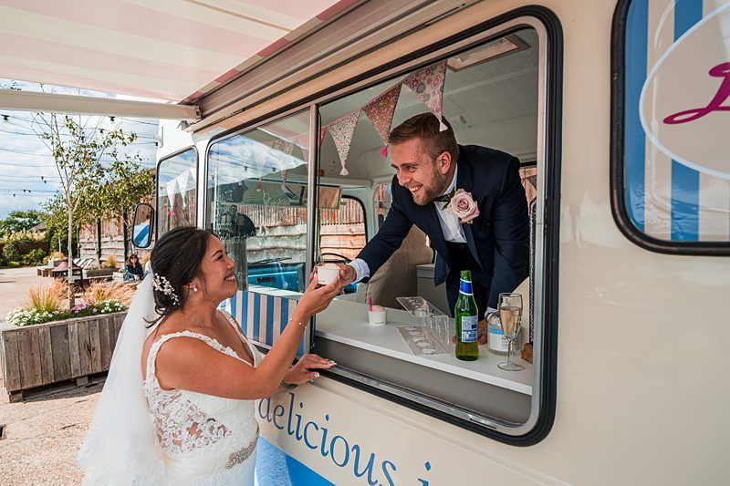 A bride in a white dress is handed an ice cream cone by a smiling man in a suit from a cream-colored ice cream van. They both appear happy and the scene is set outdoors. Image by Documentary Wedding Photographer Lewis Bishop Photography.