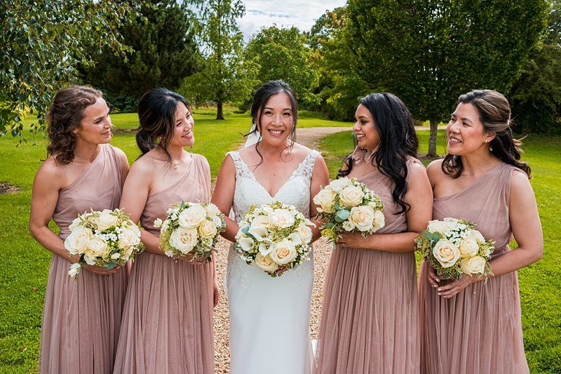A bride in a white dress holding a bouquet stands with four bridesmaids in matching light brown dresses holding similar bouquets, all smiling, in an outdoor garden setting. Image by Documentary Wedding Photographer Lewis Bishop Photography.