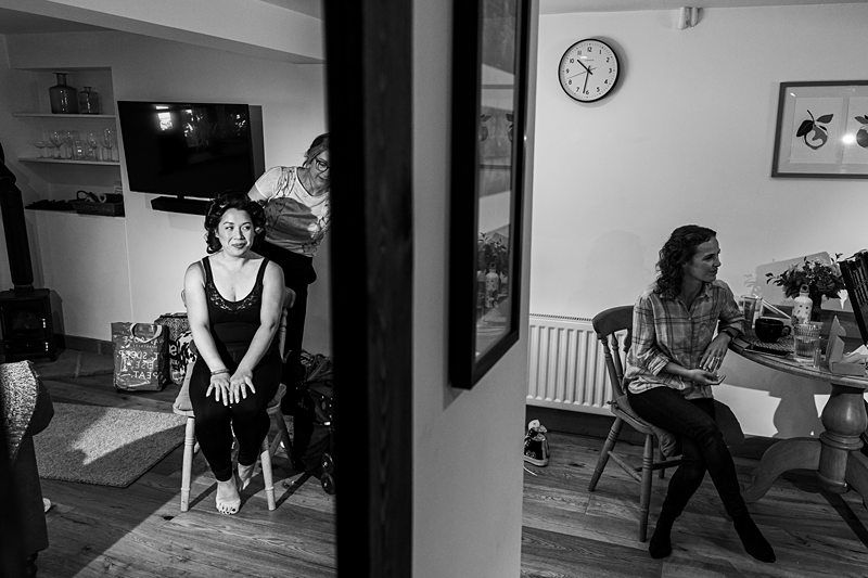 Two women are in a room; one is sitting on a chair getting her hair done, and the other is seated at a table looking away. A wall divides the room, and a clock and framed pictures hang on the walls. Image by Documentary Wedding Photographer Lewis Bishop Photography.