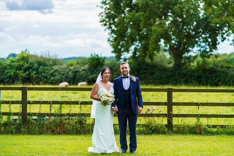 A bride and groom stand together in a grassy field in front of a wooden fence and greenery, smiling at the camera. The bride holds a bouquet of flowers. Image by Documentary Wedding Photographer Lewis Bishop Photography.