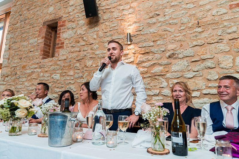A man in a white shirt is speaking into a microphone, standing at a long table set for a meal, with six seated people on either side of him against a rustic stone wall background. Image by Documentary Wedding Photographer Lewis Bishop Photography.