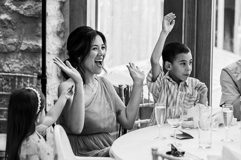 A woman and two children sit at a dining table, cheerfully raising their hands. The photo is black and white. Image by Documentary Wedding Photographer Lewis Bishop Photography.
