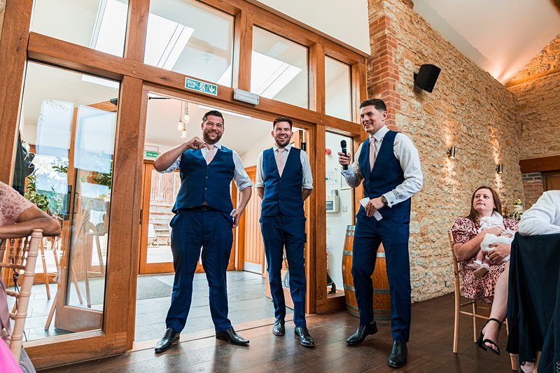 Three men in formal attire stand inside a venue with one holding a microphone. They appear to be speaking to an audience. A seated woman looks on to the right. There's a rustic brick and wooden interior. Image by Documentary Wedding Photographer Lewis Bishop Photography.