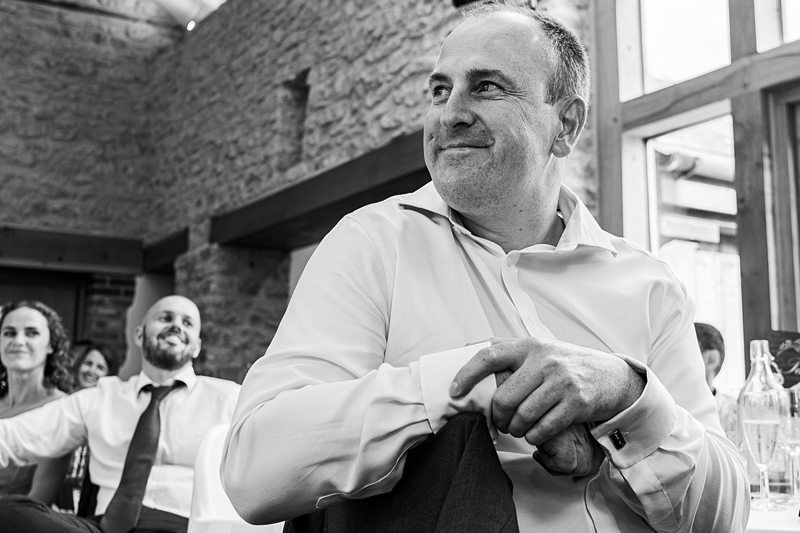 A man in a white shirt and tie sits and smiles in a rustic indoor setting with two other people visible in the background, including a man in a shirt and tie and a woman in a sleeveless dress. Image by Documentary Wedding Photographer Lewis Bishop Photography.