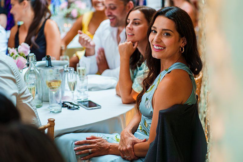 A woman in a blue dress smiles at the camera while seated at a table with others at a gathering. There are drinks and flowers on the table. Image by Documentary Wedding Photographer Lewis Bishop Photography.