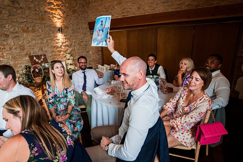 A group of people dressed in formal attire are seated around a table. One man holds up a photo, and the group looks engaged, some smiling, in a stone-walled room with wooden doors. Image by Documentary Wedding Photographer Lewis Bishop Photography.