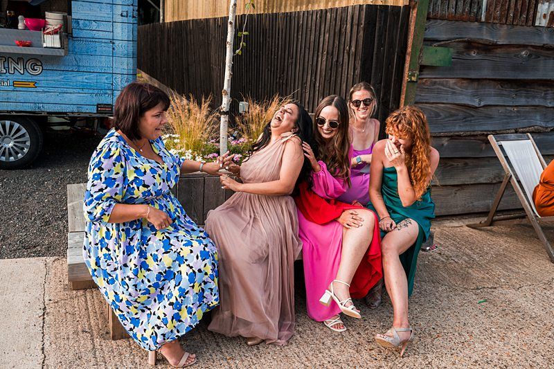 Five women sitting together on a bench outdoors, laughing and smiling. They are dressed in colorful dresses and sitting in a casual, rustic setting. Image by Documentary Wedding Photographer Lewis Bishop Photography.