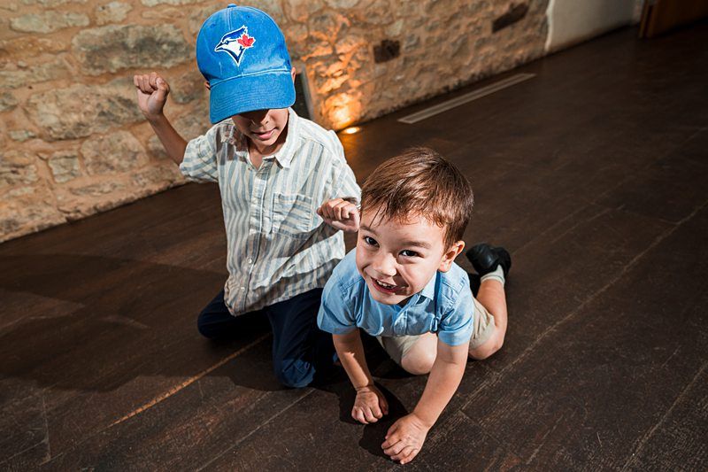 Two young boys are playing on a wooden floor, with one kneeling and smiling at the camera while the other, wearing a blue cap, poses behind him with a playful gesture. Image by Documentary Wedding Photographer Lewis Bishop Photography.