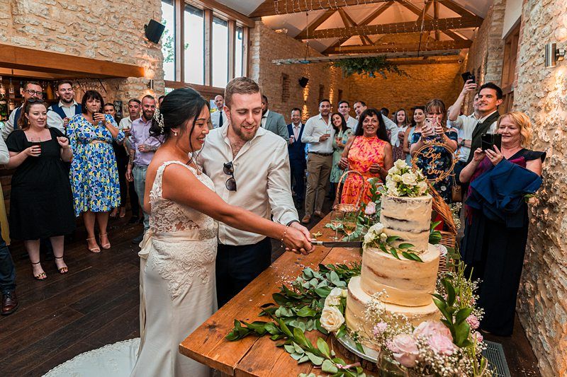 A bride and groom cut a three-tiered cake in a rustic venue. Guests are gathered around them, many taking photos. The venue features exposed brick walls and wooden beams. Image by Documentary Wedding Photographer Lewis Bishop Photography.