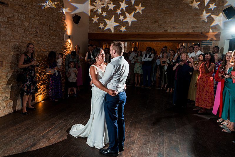 A couple shares their first dance at a wedding reception while guests watch and take photos in a warmly lit room with star-shaped lights projected on the ceiling. Image by Documentary Wedding Photographer Lewis Bishop Photography.