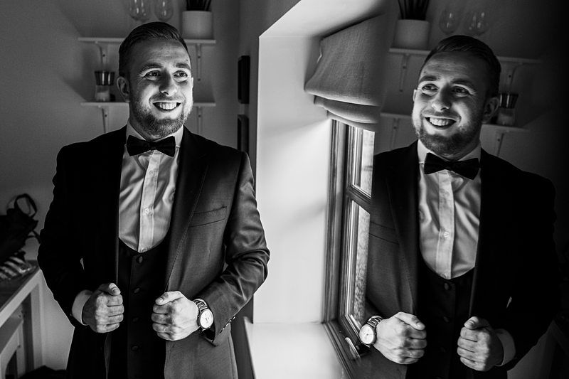 Black and white image of a smiling man in a suit with a bow tie, standing in front of a mirror, adjusting his jacket. Image by Documentary Wedding Photographer Lewis Bishop Photography.