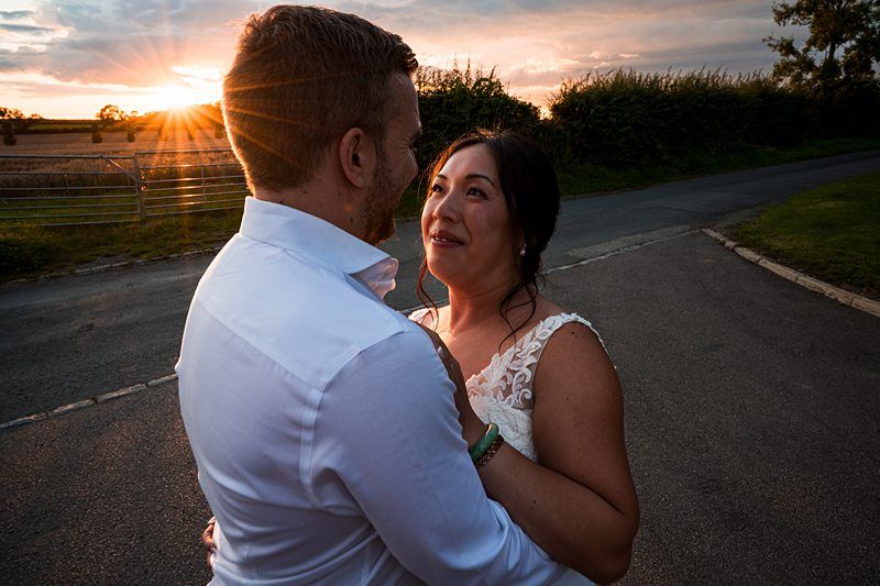 A couple in wedding attire gazes at each other lovingly while standing on a paved road during sunset with a field and hedgerow in the background. Image by Documentary Wedding Photographer Lewis Bishop Photography.