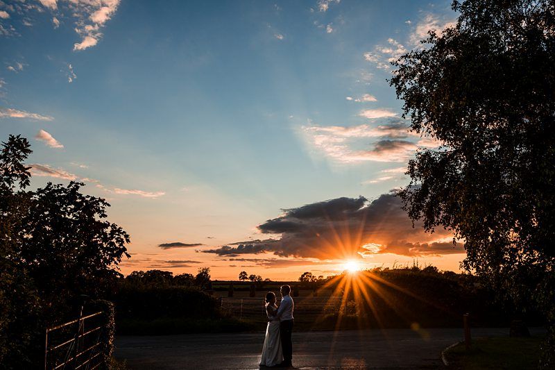 A couple stands close together under a vibrant sunset sky, with the sun partially obscured by clouds, casting rays of light across a rural landscape. Trees frame the scene. Image by Documentary Wedding Photographer Lewis Bishop Photography.