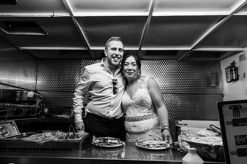A couple in formal attire, with the woman in a wedding dress and the man in a shirt and trousers, stand smiling behind a counter with food trays in a kitchen. Image by Documentary Wedding Photographer Lewis Bishop Photography.