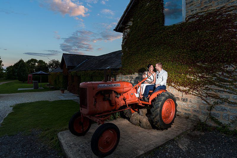 A couple sits on an old, red Allis-Chalmers tractor parked beside a vine-covered stone building at dusk. Image by Documentary Wedding Photographer Lewis Bishop Photography.