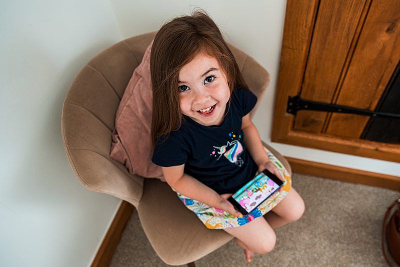 A young girl with long brown hair sitting on a chair and holding a smartphone, looking up and smiling. She is wearing a dark t-shirt with a colorful design and a patterned skirt. Image by Documentary Wedding Photographer Lewis Bishop Photography.