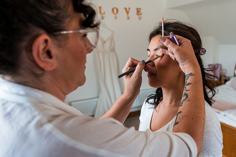 A makeup artist applies eye makeup to a woman in a white shirt in a room with a wedding dress hanging on a wall that spells "LOVE. Image by Documentary Wedding Photographer Lewis Bishop Photography.