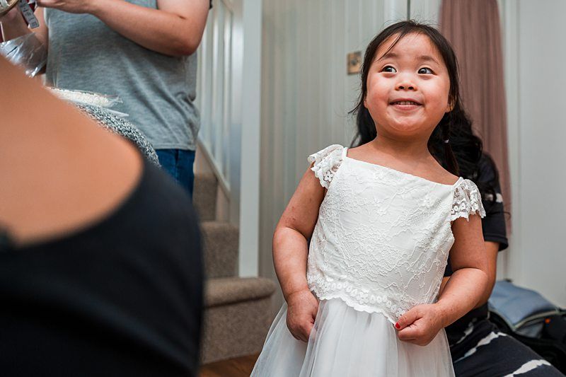 Young child in a white dress smiling, standing indoors near a staircase with an adult partially visible in the background. Image by Documentary Wedding Photographer Lewis Bishop Photography.