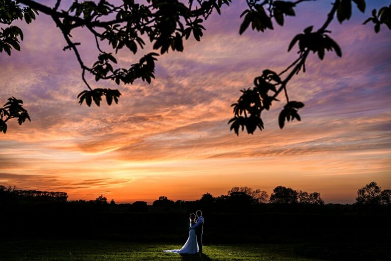 A couple stands in an open field, embracing at sunset with dramatic, colorful clouds overhead and silhouetted tree branches framing the scene. Image by Documentary Wedding Photographer Lewis Bishop Photography.