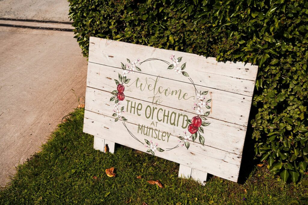 A weathered wooden sign with floral accents leans against a hedge. The sign reads "Welcome to The Orchard at Munstey" and features painted roses. The sign rests on a grassy area next to a concrete path. Image by Documentary Wedding Photographer Lewis Bishop Photography.