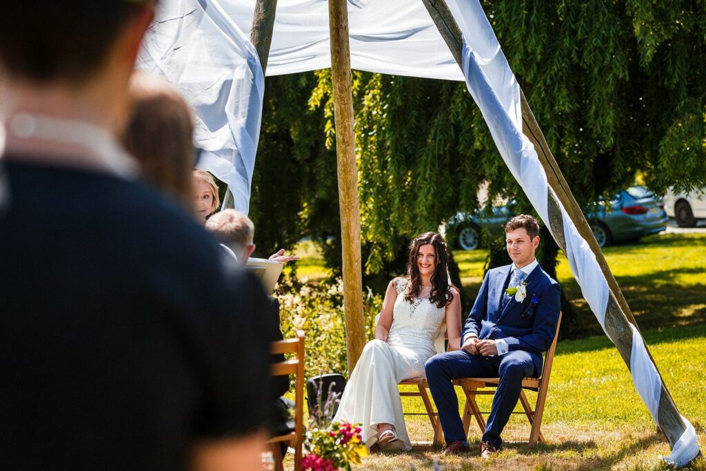 A bride in a white dress and a groom in a blue suit sit under a white canopy during an outdoor wedding ceremony. They are seated on wooden chairs with greenery and a few parked cars in the background. Partially blurred guests are visible in the foreground. Image by Documentary Wedding Photographer Lewis Bishop Photography.