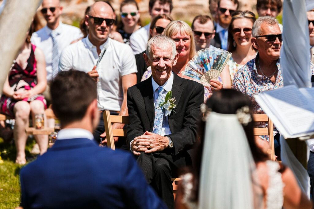 An elderly man in a suit and tie, smiling and seated outdoors, surrounded by a crowd of casually dressed people. The man is facing a couple in wedding attire, who are turned away from the camera. The setting appears to be a sunny, outdoor wedding ceremony. Image by Documentary Wedding Photographer Lewis Bishop Photography.