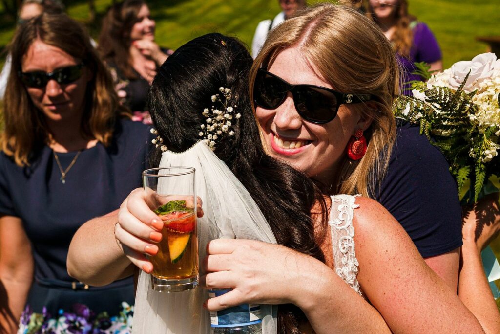 A bride in a white veil and flower hairpiece is hugging a woman wearing sunglasses and large earrings. The woman is holding a drink garnished with a strawberry and mint. Several people in the background are smiling and enjoying the outdoor event. Image by Documentary Wedding Photographer Lewis Bishop Photography.