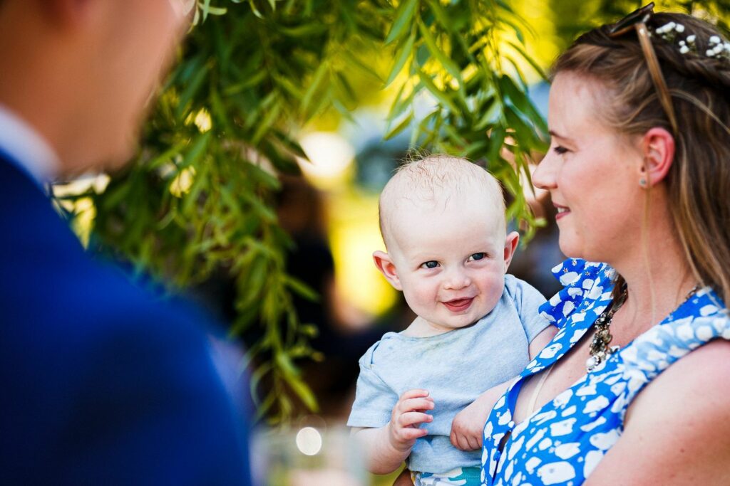 A smiling woman in a blue patterned dress is holding a cheerful baby in her arms. They are outdoors, and the background is softly blurred with green foliage. An out-of-focus figure in a blue suit is in the foreground. The baby is reaching out towards the figure. Image by Documentary Wedding Photographer Lewis Bishop Photography.