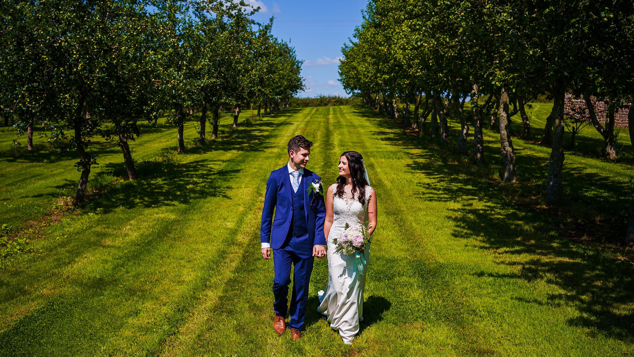 A bride and groom walk hand in hand through a lush, green orchard on a sunny day. The groom is in a blue suit with a white boutonniere, and the bride wears a white wedding dress, holding a bouquet of flowers. Trees line both sides of the path they walk on. Image by Documentary Wedding Photographer Lewis Bishop Photography.