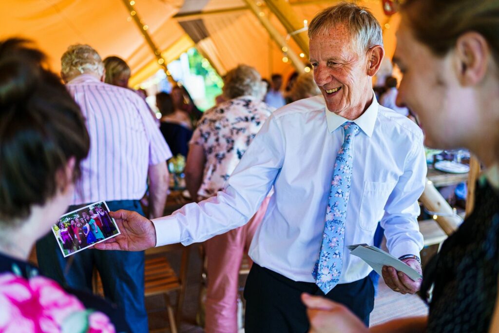 An elderly man in a white shirt and patterned tie is smiling while showing a photograph to two women at a lively indoor gathering. Decorated with warm lighting, the setting appears festive with people mingling in the background. Image by Documentary Wedding Photographer Lewis Bishop Photography.