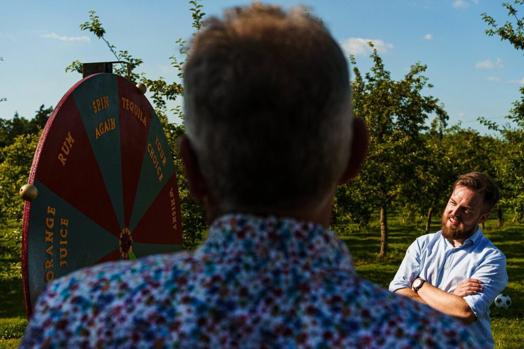 Two men stand outdoors by a large spinning wheel with colorful segments labeled with drink options like "Rum" and "Tequila." One man, seen from behind, watches the wheel while the other, facing the wheel, stands with his arms folded, smiling slightly. Trees fill the background. Image by Documentary Wedding Photographer Lewis Bishop Photography.