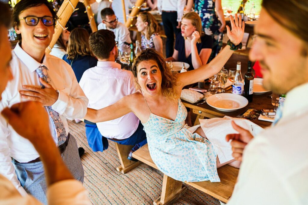 A joyful woman in a light blue floral dress sits on a bench with her arms spread wide, laughing enthusiastically at a lively gathering. People around her are engaged in conversation and celebration, creating a festive atmosphere. Image by Documentary Wedding Photographer Lewis Bishop Photography.