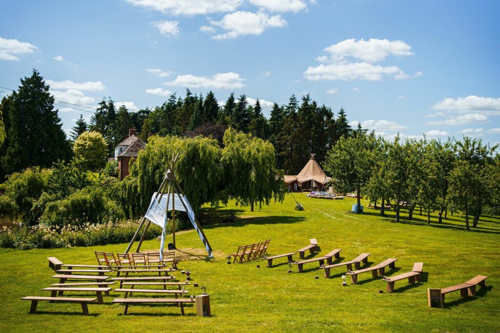 A picturesque outdoor wedding setup on a sunny day, with wooden benches arranged in arcs around a small arch made of tree branches and a white cloth. A tipi-style tent is seen in the background, surrounded by green grass, trees, and a clear blue sky. Image by Documentary Wedding Photographer Lewis Bishop Photography.