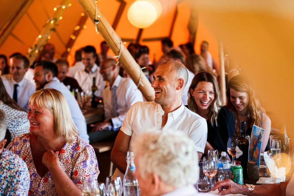 A lively group of people at a social gathering, seated closely around tables decorated with wine glasses and bottles. The scene is warmly lit, with fairy lights adding ambiance. Everyone is smiling and laughing, creating a joyful and celebratory atmosphere. Image by Documentary Wedding Photographer Lewis Bishop Photography.