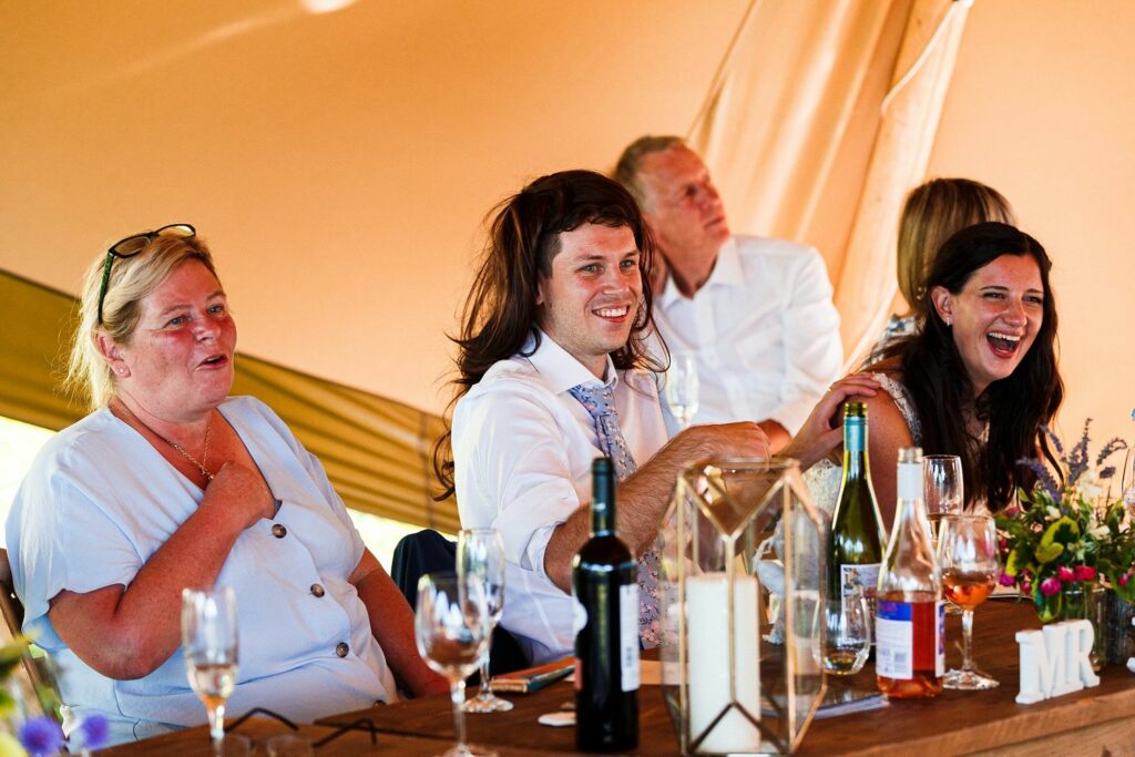 A group of people sits at a wooden table during a wedding reception. Some laugh while one man wears a wig. Bottles of wine and glasses cover the table. A "MR" sign is visible next to decorative lanterns and flower arrangements. Everyone appears to be enjoying the event. Image by Documentary Wedding Photographer Lewis Bishop Photography.