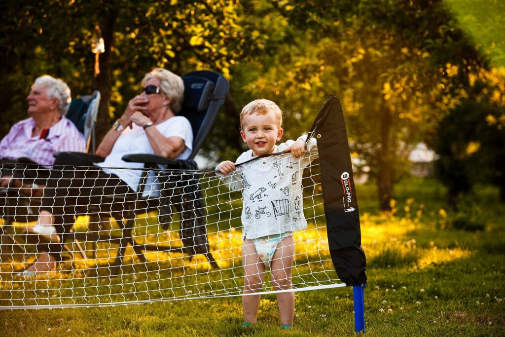 A small child in a diaper and shirt stands by a badminton net outdoors, smiling at the camera. Two elderly women sit in reclining chairs in the background, also smiling. The scene is lit by warm, golden sunlight, suggesting late afternoon or early evening. Image by Documentary Wedding Photographer Lewis Bishop Photography.