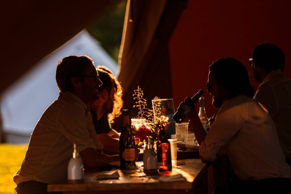 Several people are gathered around a wooden table at an outdoor event during sunset. They are engaged in conversation, with some holding drinks. The warm glow of the setting sun illuminates the scene, creating a cozy atmosphere. In the foreground, a vase with flowers is visible. Image by Documentary Wedding Photographer Lewis Bishop Photography.