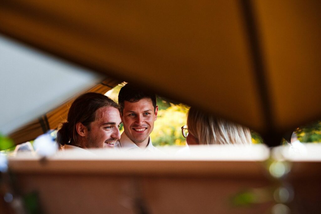 Three people are smiling and conversing under an outdoor canopy. The image is framed between the edges of the canopy, creating a warm and inviting atmosphere with natural greenery in the background. The focus is on the joyful expressions of the people. Image by Documentary Wedding Photographer Lewis Bishop Photography.