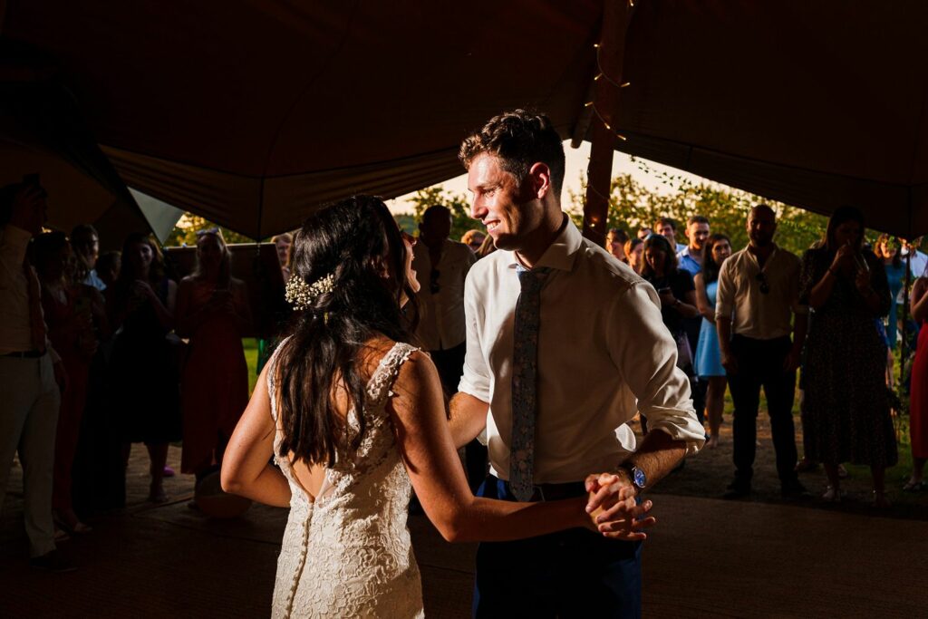 A newlywed couple shares a dance under a canopy tent at sunset. The groom, in a white shirt and blue tie, gazes lovingly at the bride, who is wearing a lace wedding dress with floral hair accessories. Guests stand around them, watching and taking pictures. Image by Documentary Wedding Photographer Lewis Bishop Photography.