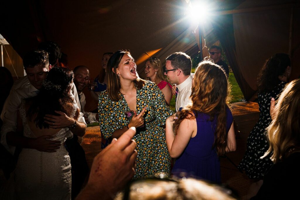 A group of people are joyfully dancing at what appears to be a nighttime event or celebration, with one woman in the center singing along passionately. The scene is lively with a bright spotlight and warm ambient lighting. Image by Documentary Wedding Photographer Lewis Bishop Photography.