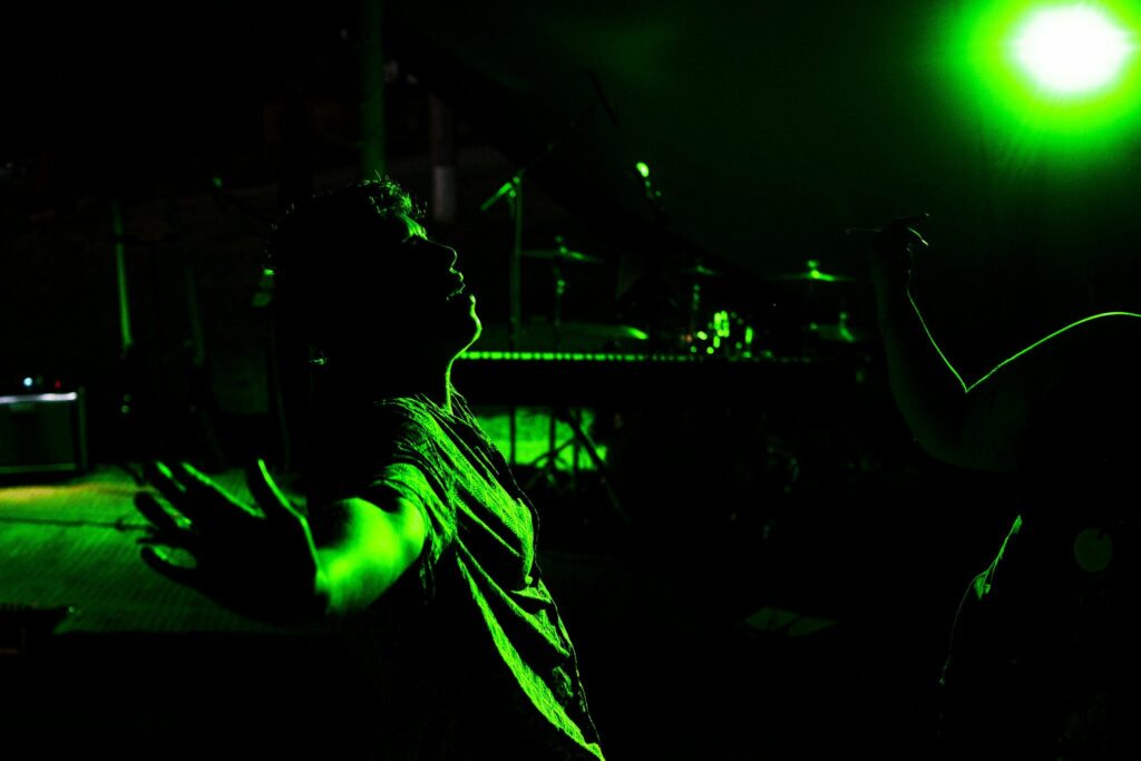 A person stands with arms outstretched, bathed in green stage lighting. The background features musical instruments, including a keyboard and drum set, suggesting a live music performance or a concert setting. Another person is partially visible with a raised hand. Image by Documentary Wedding Photographer Lewis Bishop Photography.