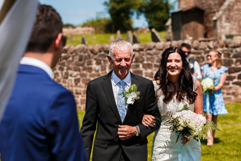 A bride in a white dress and a man in a black suit walk arm-in-arm down an outdoor aisle. The man holds a small bouquet and smiles. They are walking towards a person in a blue suit. Several people are visible in the background, and it is a sunny day. Image by Documentary Wedding Photographer Lewis Bishop Photography.