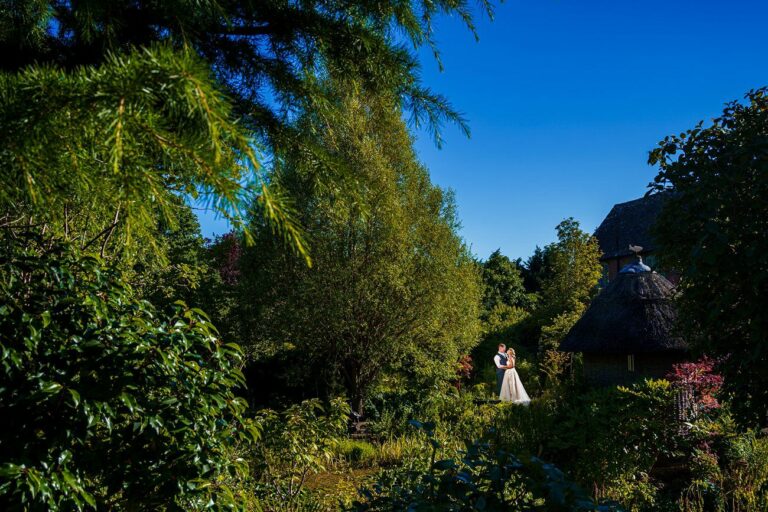 A couple in wedding attire stands close together in an open area surrounded by lush greenery and trees under a clear blue sky. Nearby is a thatched-roof structure partially hidden by the foliage. The scene has a serene, romantic atmosphere. Image by Documentary Wedding Photographer Lewis Bishop Photography.