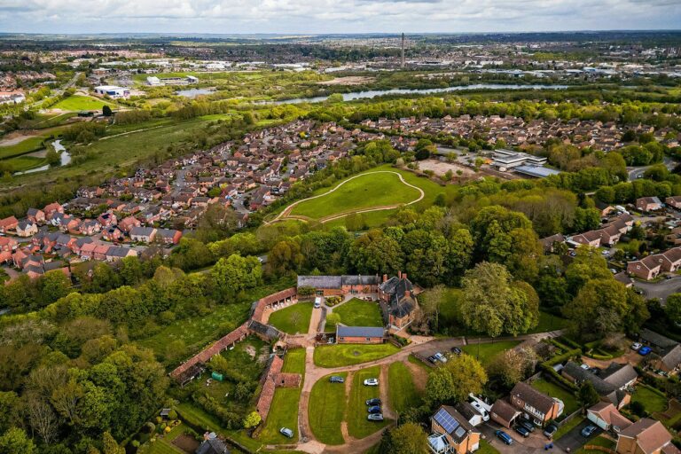 Aerial view of a residential area with houses, green spaces, and a castle-like structure surrounded by trees. A larger town and expansive landscape can be seen in the background. Image by Documentary Wedding Photographer Lewis Bishop Photography.
