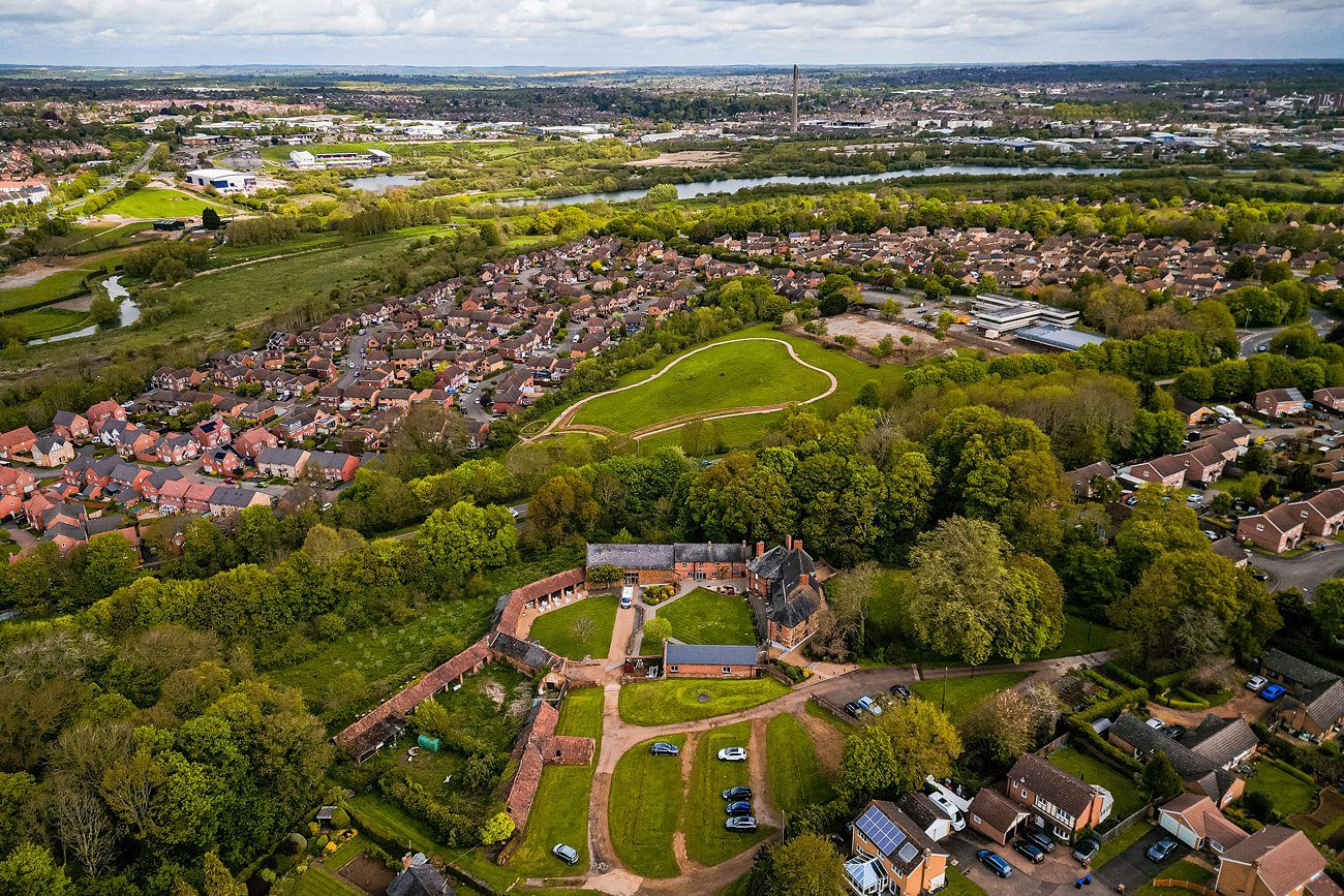 Aerial view of a residential area with houses, green spaces, and a castle-like structure surrounded by trees. A larger town and expansive landscape can be seen in the background. Image by Documentary Wedding Photographer Lewis Bishop Photography.